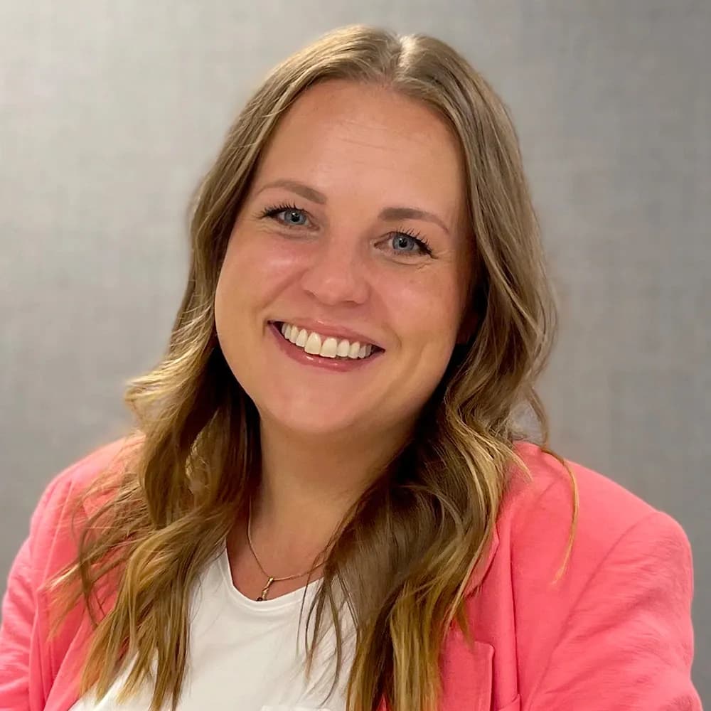 Smiling woman with wavy hair, wearing a pink blazer and white shirt, against a gray background.