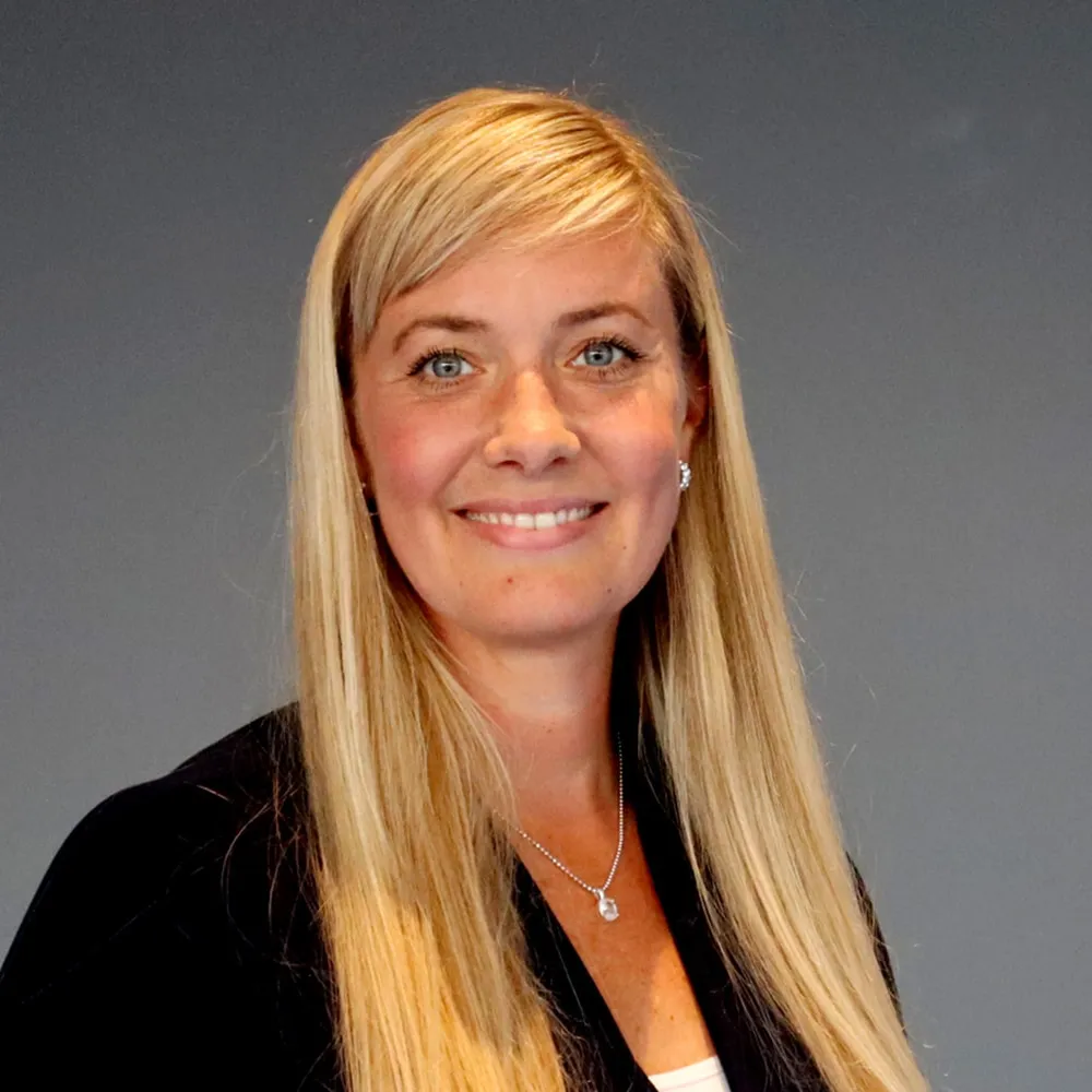 Smiling person with long blond hair, wearing a dark jacket and light top, against a plain gray background.