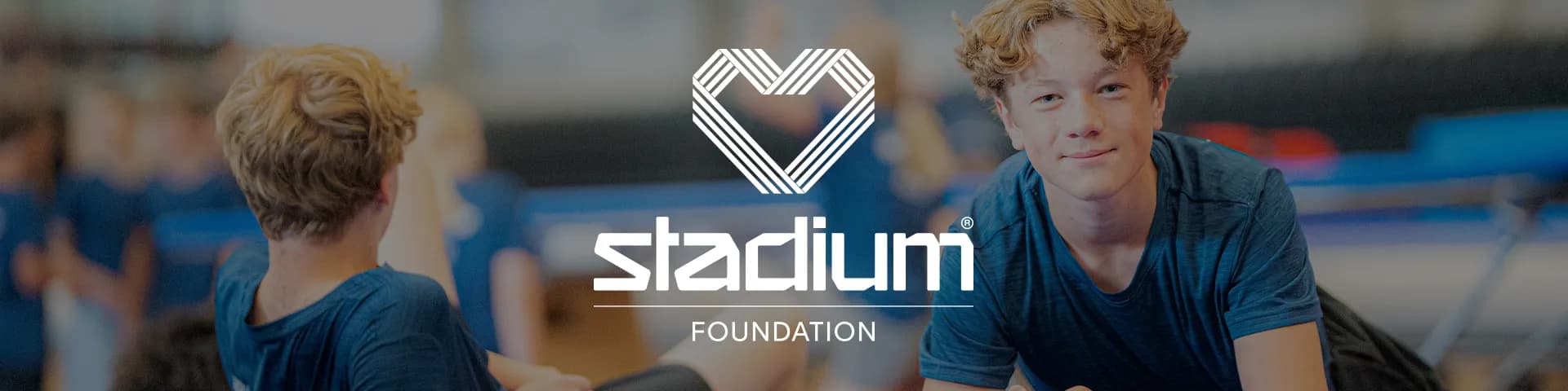 Two boys in blue shirts lying on gym mats with a heart logo and "Stadium Foundation" text overlaid. Background shows a blurred gym setting.