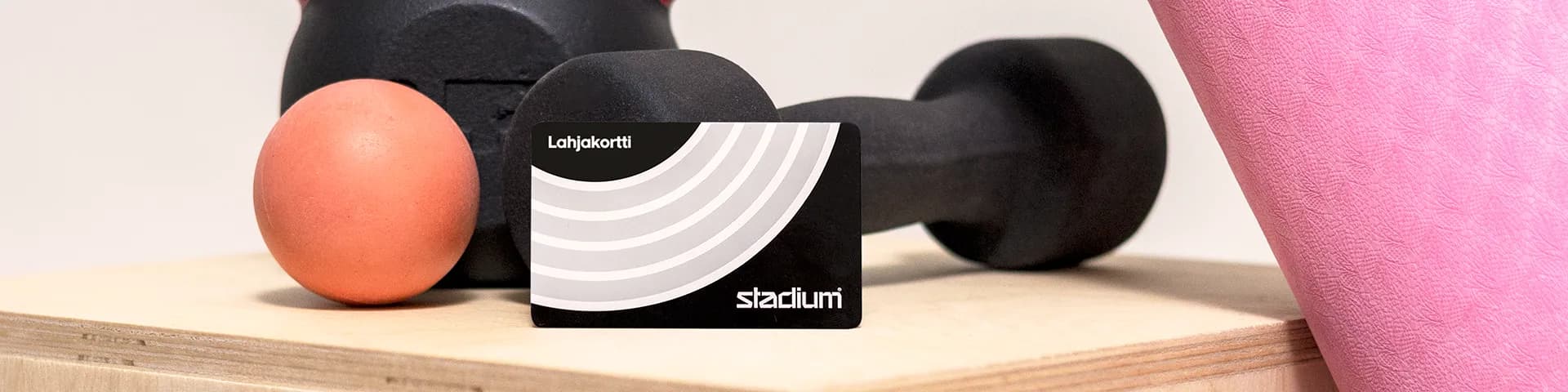 A black card labeled "Leikjakort" leans against a dumbbell on a wooden surface, with an orange ball and a pink mat nearby.