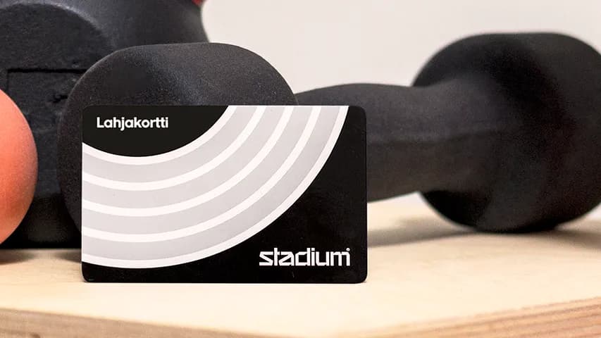 A black card labeled "Leikjakort" leans against a dumbbell on a wooden surface, with an orange ball and a pink mat nearby.