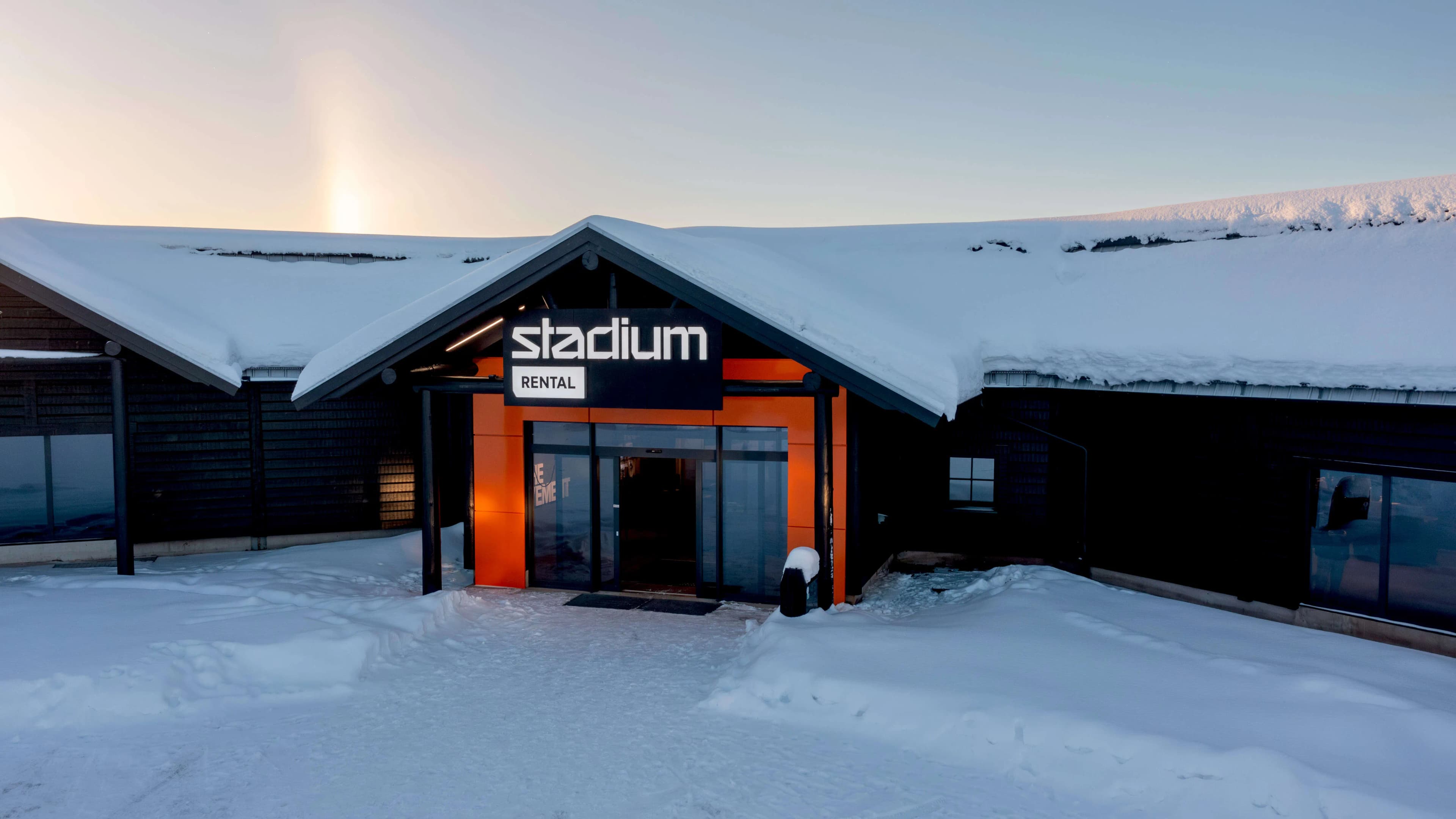 A snow-covered building with a glass entrance and "Stadium Rental" sign, under a clear sky with a faint sun.