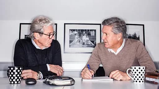 Two men sitting at a table with black-and-white mugs, discussing and writing, in front of framed photos on the wall.