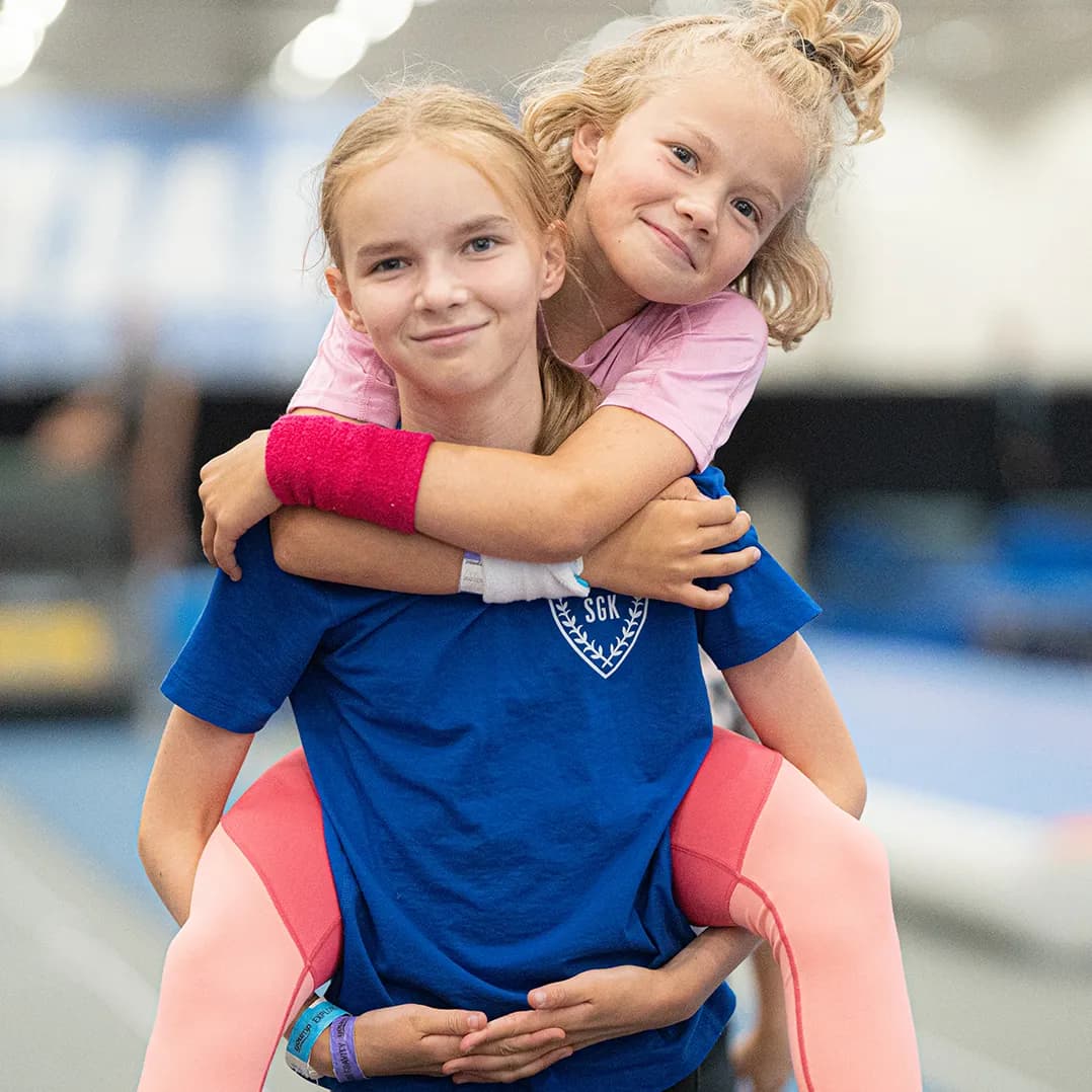Two smiling girls in athletic wear, one giving the other a piggyback ride in a gym. The girl in blue holds the other securely.