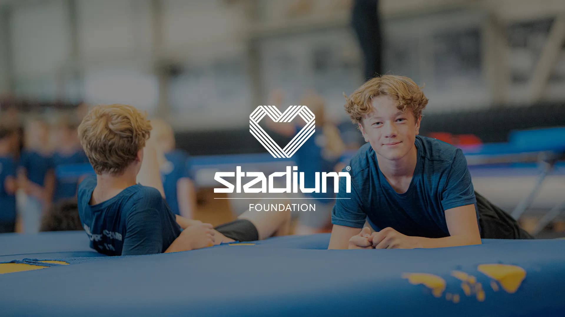 Two boys in blue shirts lying on gym mats with a heart logo and "Stadium Foundation" text overlaid. Background shows a blurred gym setting.